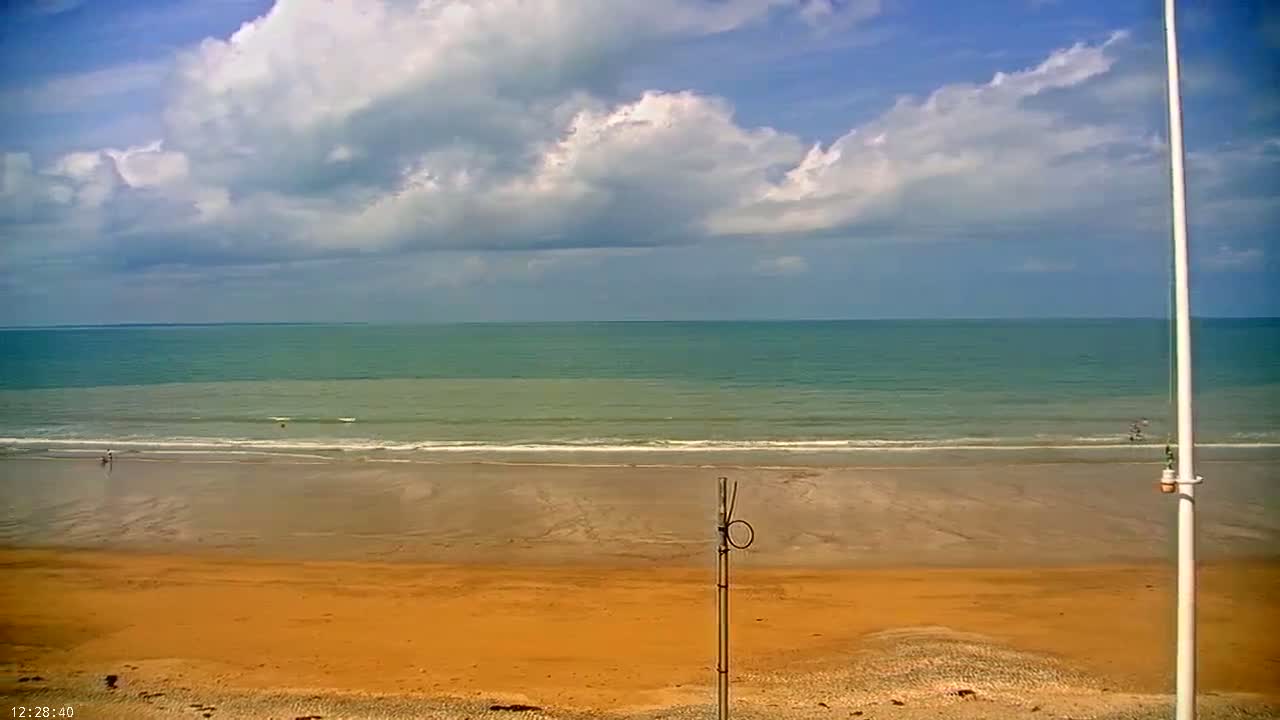 A mostly sunny day shows a wide sandy beach with a calm ocean and a few people visible in the distance.
