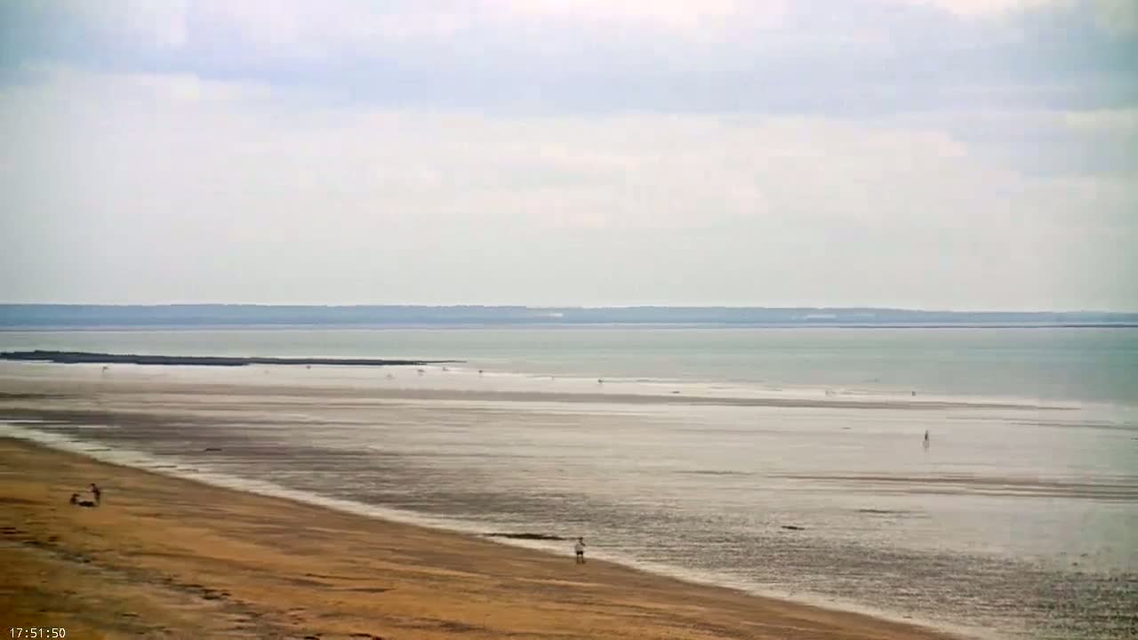 A wide expanse of sandy beach and calm, shallow water stretches to a distant shoreline under a cloudy sky.