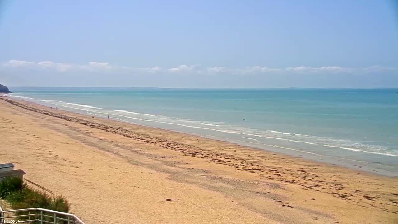 A long, sandy beach stretches toward a calm, light-blue ocean under a mostly sunny sky with a few scattered clouds.