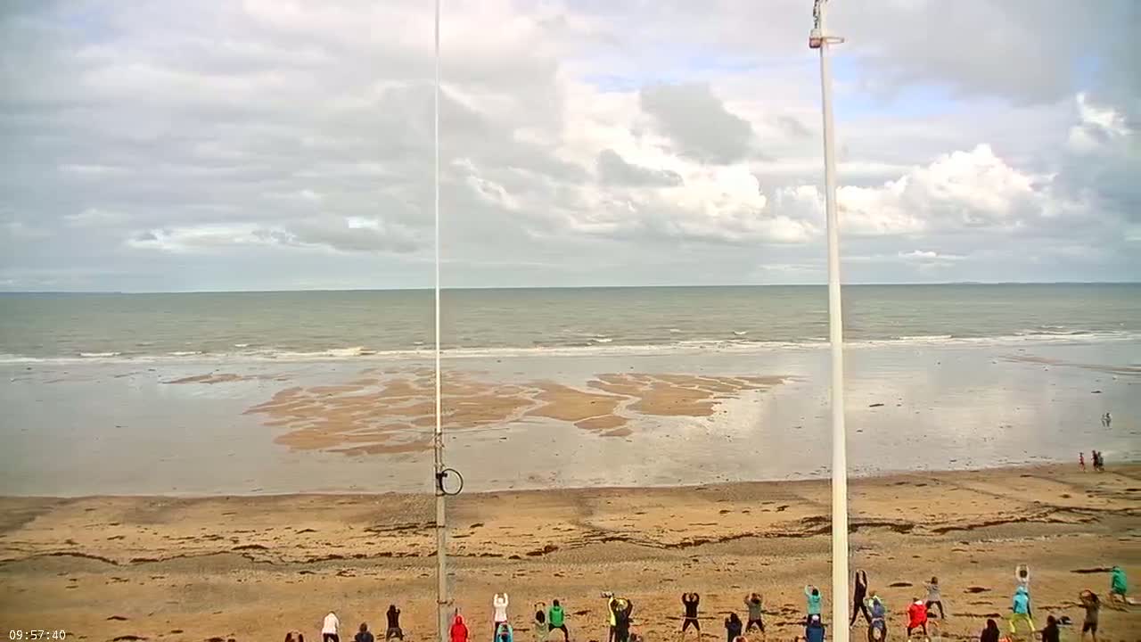 A group of people are exercising on a sandy beach under a partly cloudy sky, with calm ocean waters in the background.