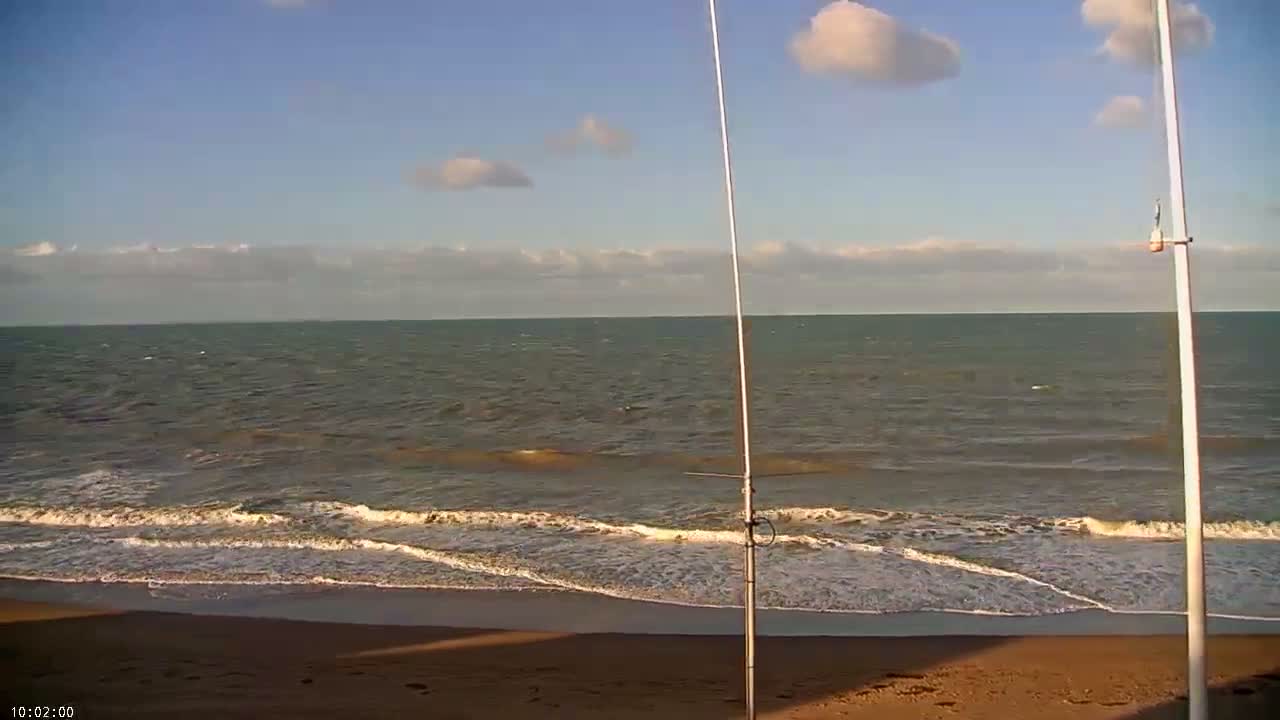 A bright, partly cloudy sky hangs over a vast, choppy ocean with waves breaking on a sandy beach, featuring two slender white poles on the shore.