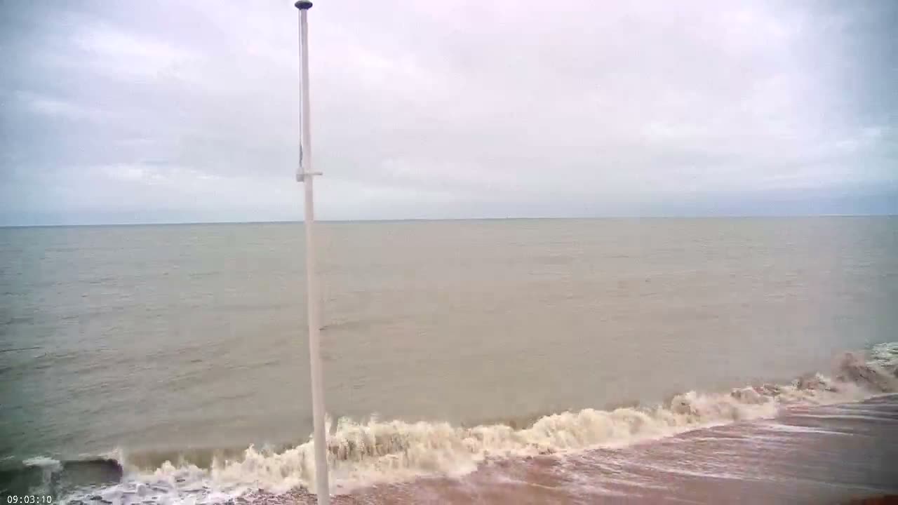 A vast grey ocean under an overcast sky has choppy waves breaking on a pebble beach, with a tall white pole standing prominently in the foreground.