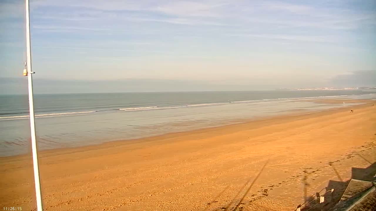 A vast grey ocean under an overcast sky has choppy waves breaking on a pebble beach, with a tall white pole standing prominently in the foreground.