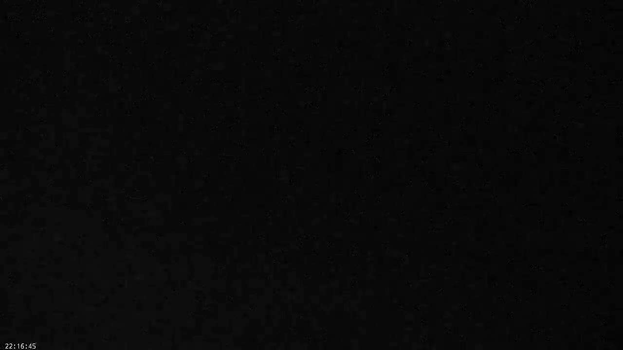 A vast grey ocean under an overcast sky has choppy waves breaking on a pebble beach, with a tall white pole standing prominently in the foreground.