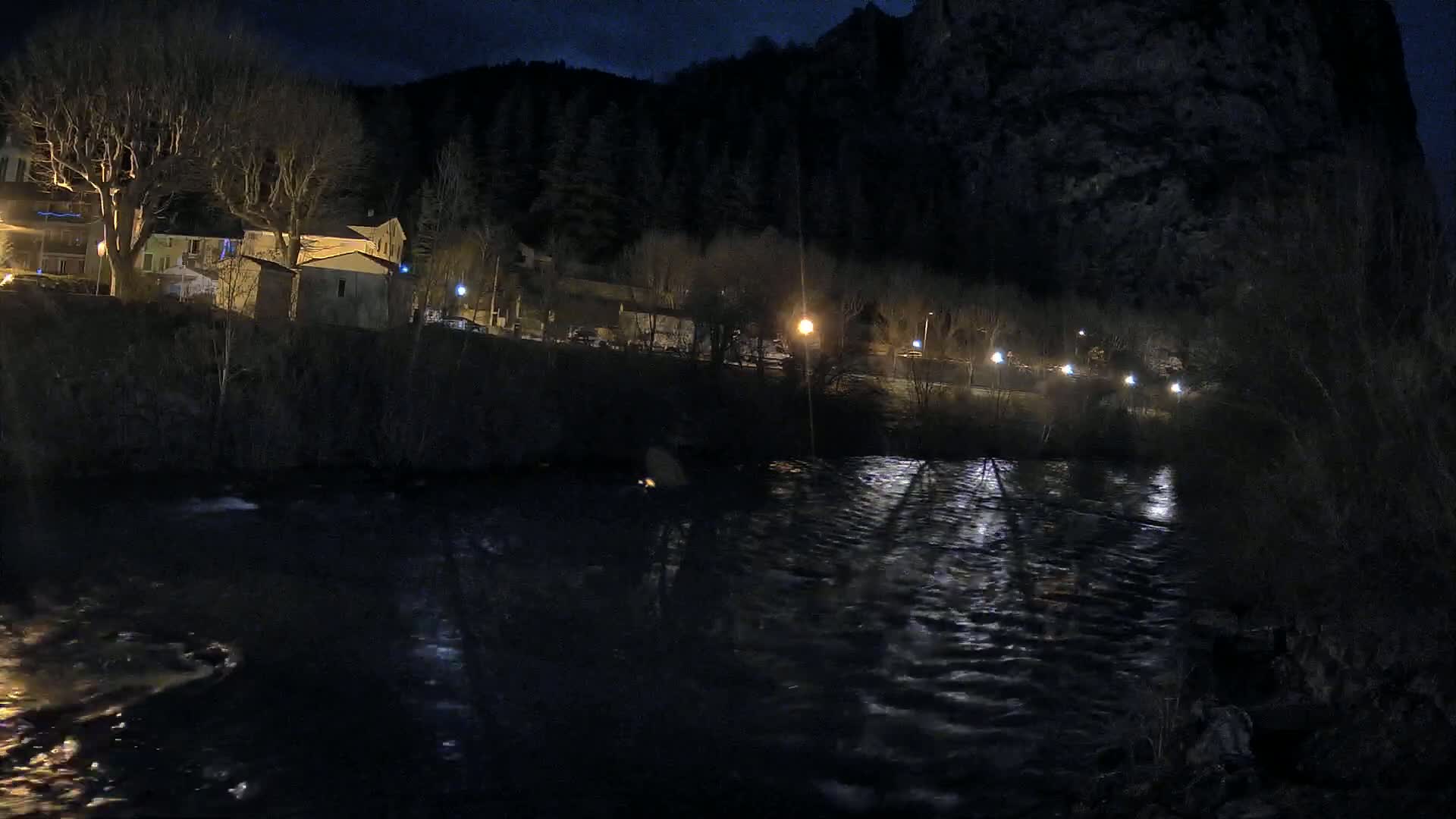 A dark, nighttime scene shows a flowing river reflecting lights from a village nestled beside a rocky hillside under a clear, moonlit sky.
