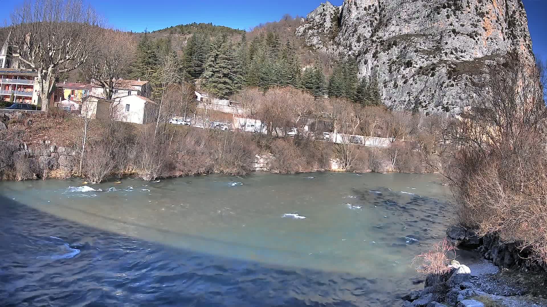 A river flows past a village nestled beside a steep, rocky cliff under a sunny sky.