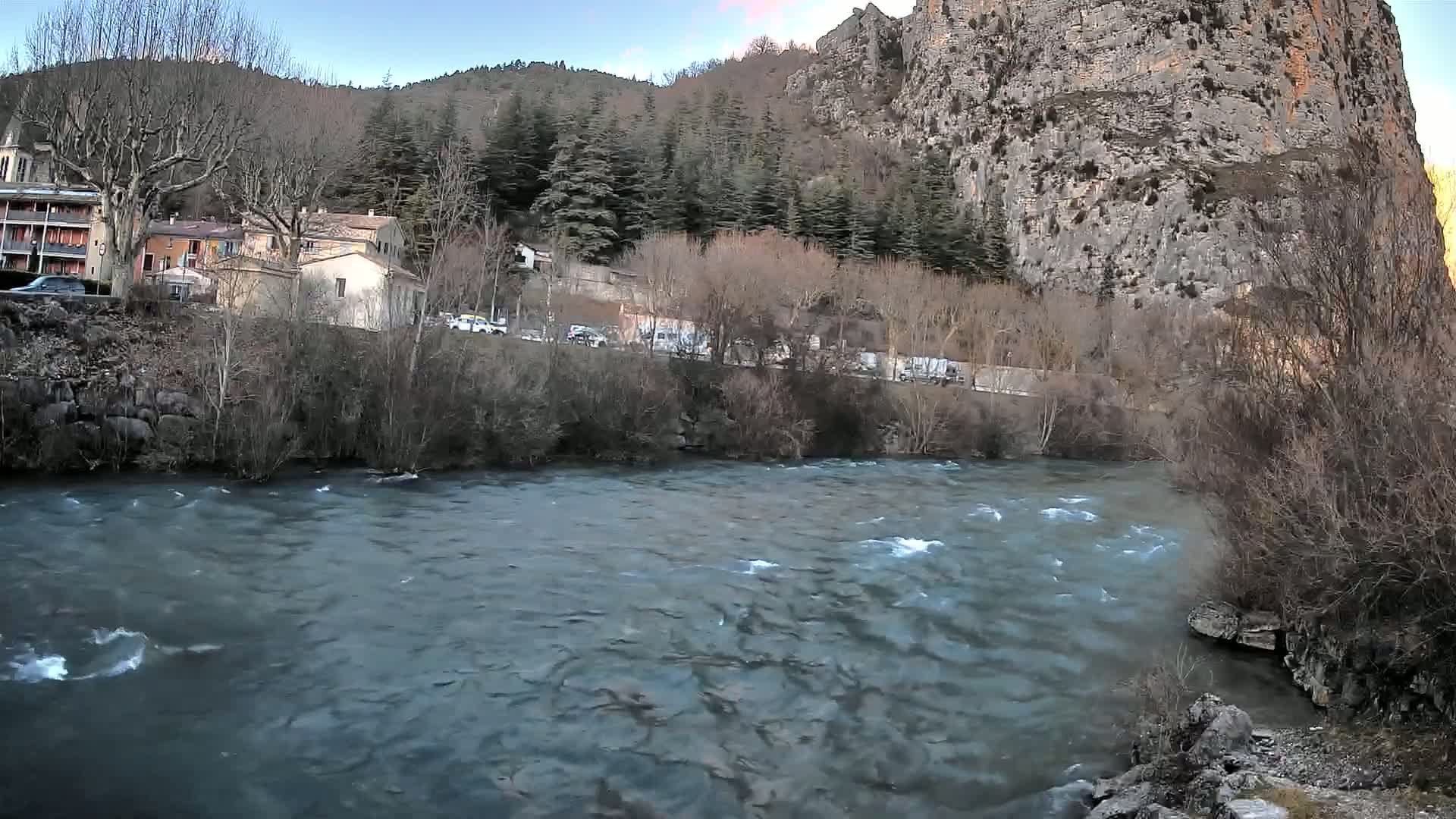 A swiftly flowing river runs in front of a village nestled beside a steep, rocky cliff and a forest of mostly bare winter trees under a mostly clear sky.