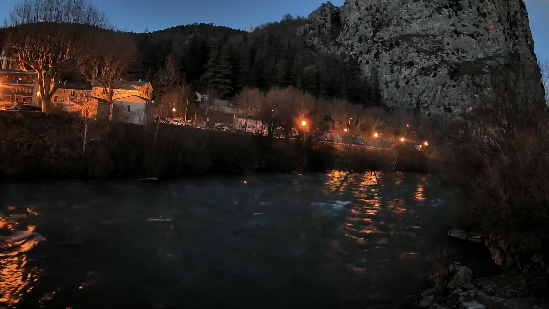 A river flows past a village at twilight, under a large cliff, illuminated by streetlights.