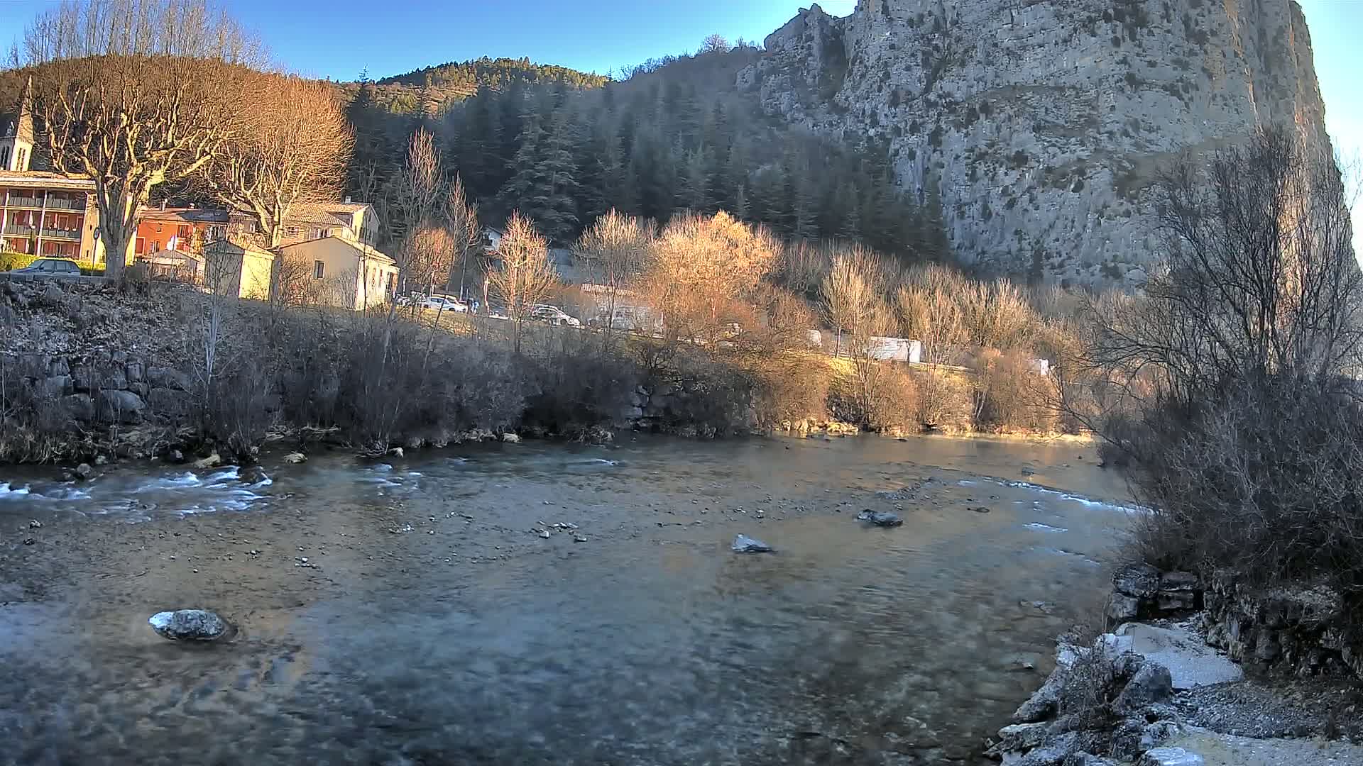 A slow-moving river flows past a village nestled at the base of a large, light-grey cliff under a clear blue sky.