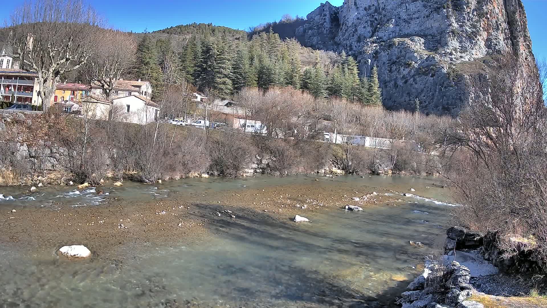 A shallow, rocky river flows past a village nestled beneath a steep, rocky cliff and a forest of evergreens, under a sunny sky.