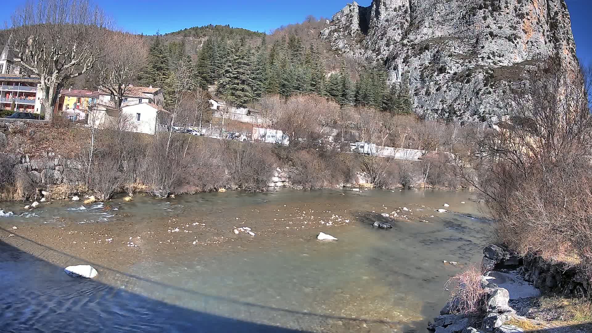 A slow-moving river flows past a village nestled beside a large, rocky cliff face under a clear, sunny sky.