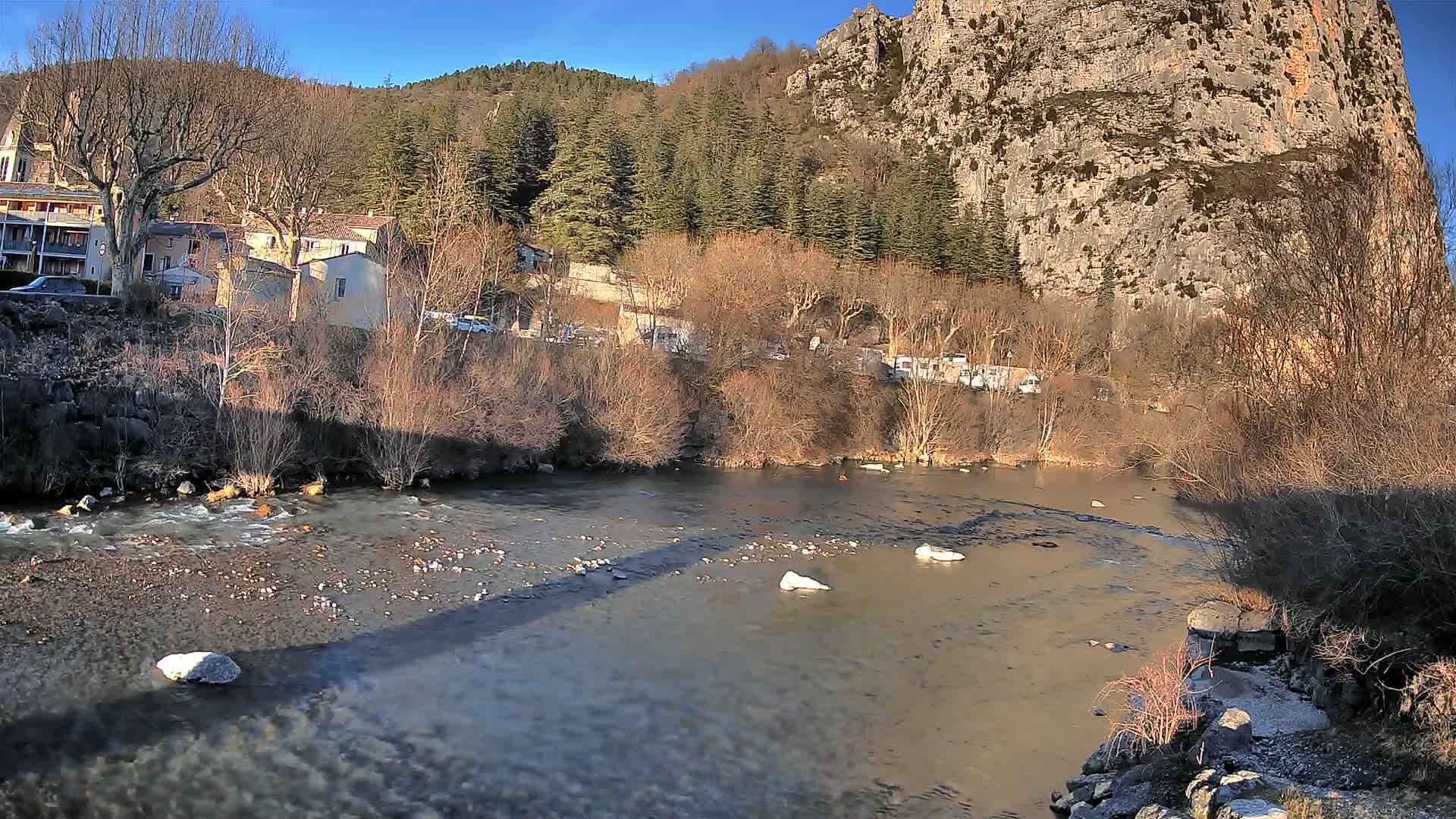 A slow-moving river flows past a village nestled at the base of a large, rocky cliff under a clear, sunny sky.