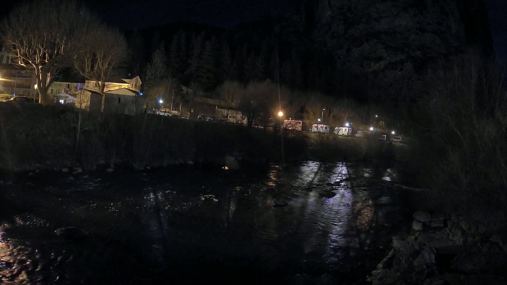 A dark, nighttime scene shows a river flowing past buildings and trees illuminated by sparse streetlights.