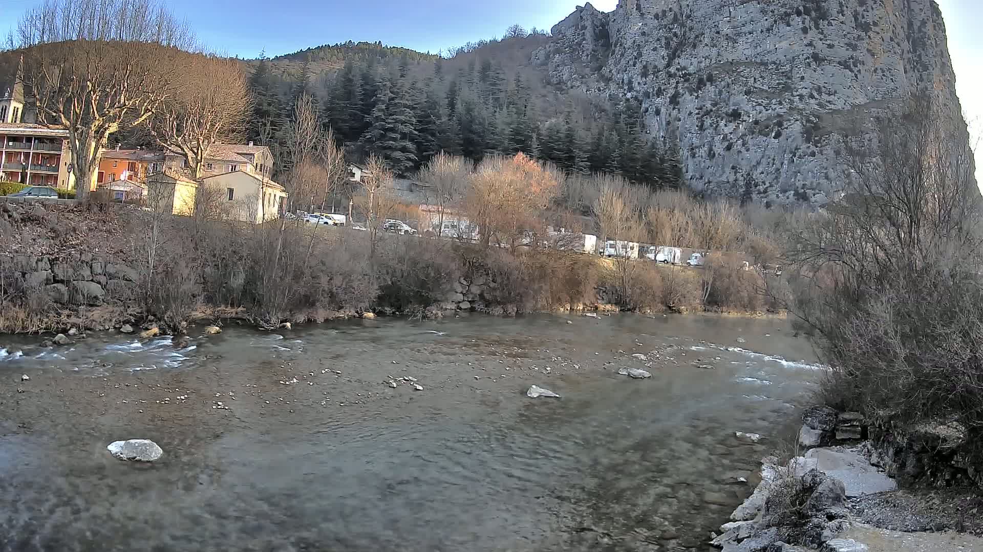 A clear river flows past a village nestled at the base of a large, rocky cliff face under a mostly clear sky.