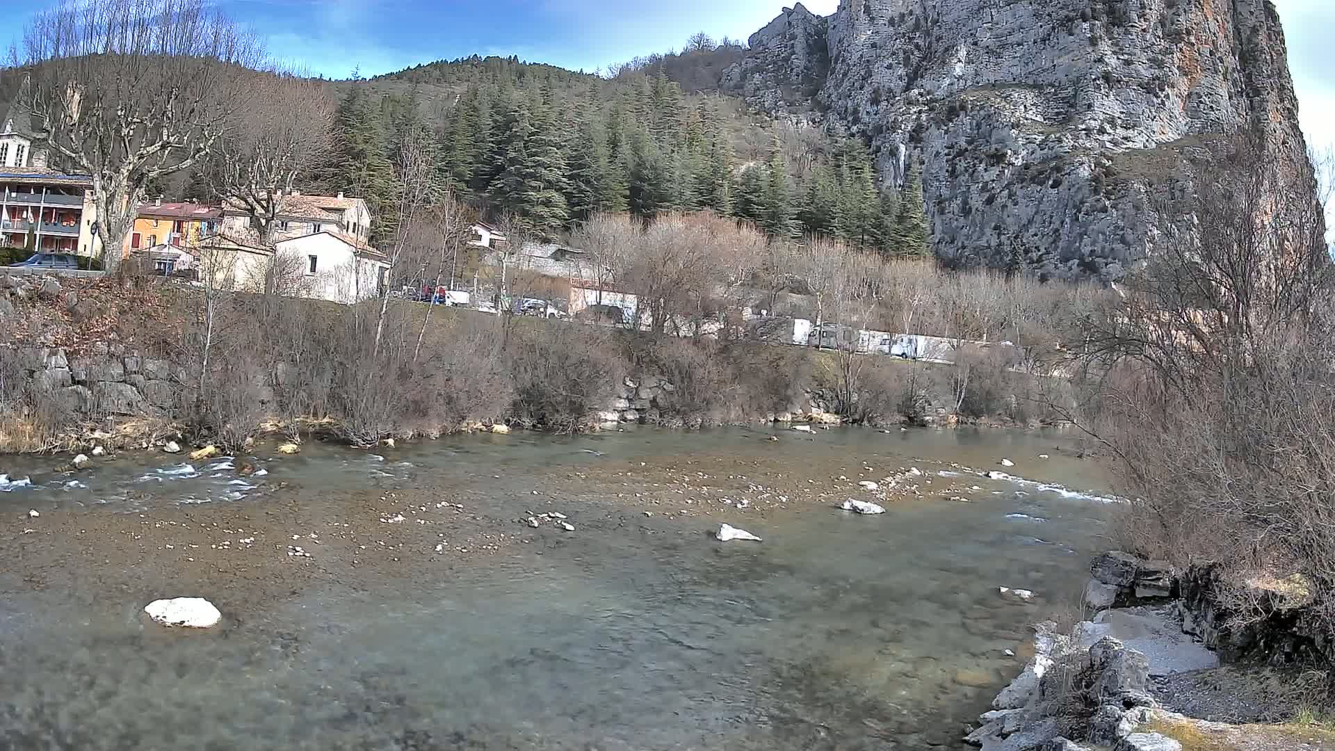 A clear, shallow river flows past a village nestled beside a steep, rocky cliff under a bright, sunny sky.