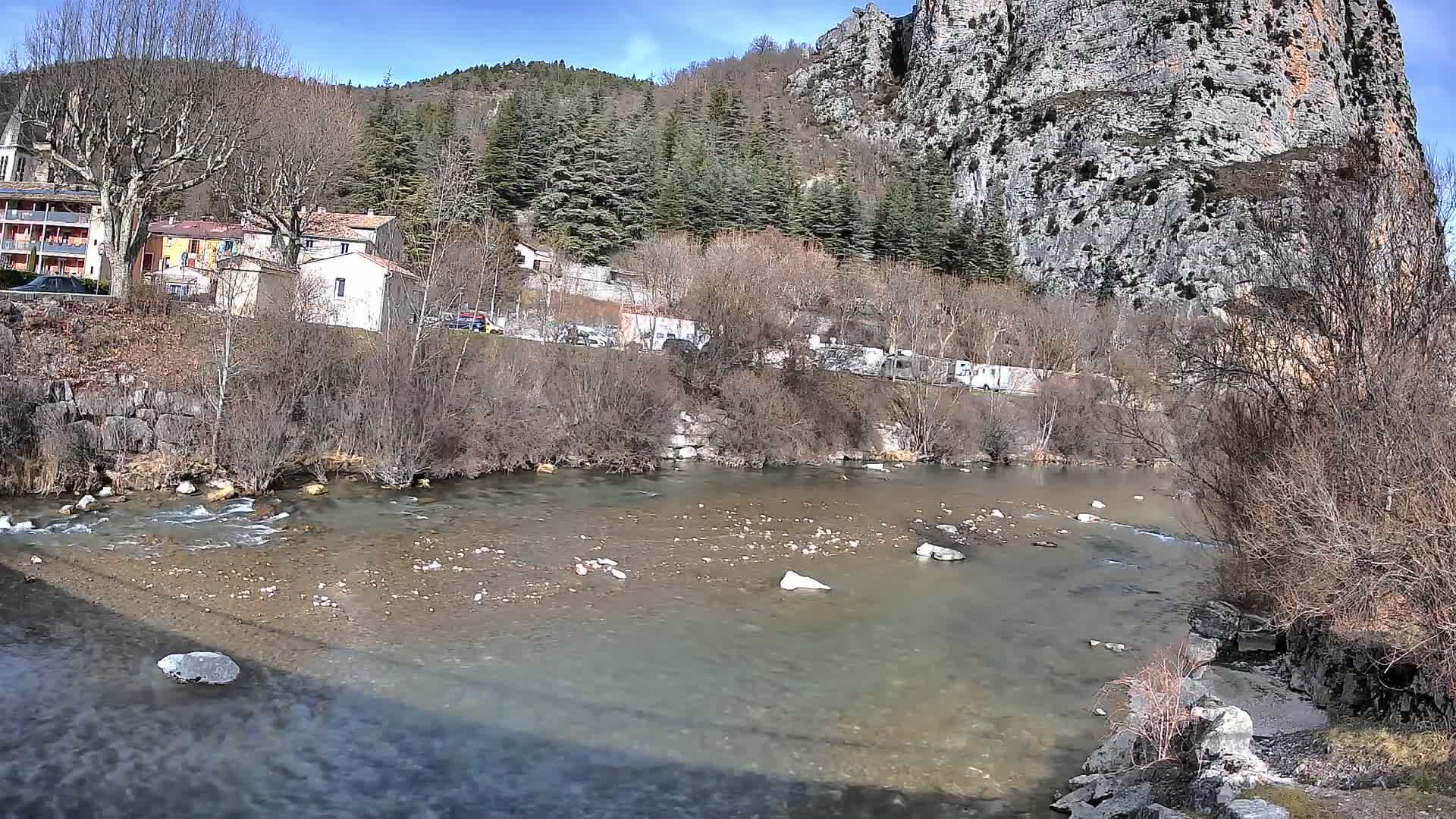 A clear, shallow river flows past a village nestled in a valley alongside a large, rocky cliff face under a sunny sky.