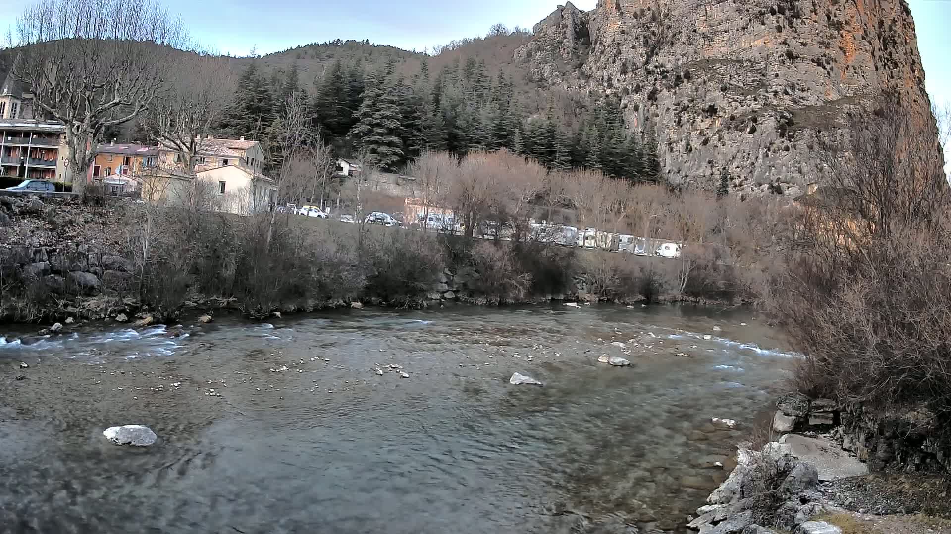 A clear river flows past a village nestled beside a large, rocky cliff under a cloudy sky.