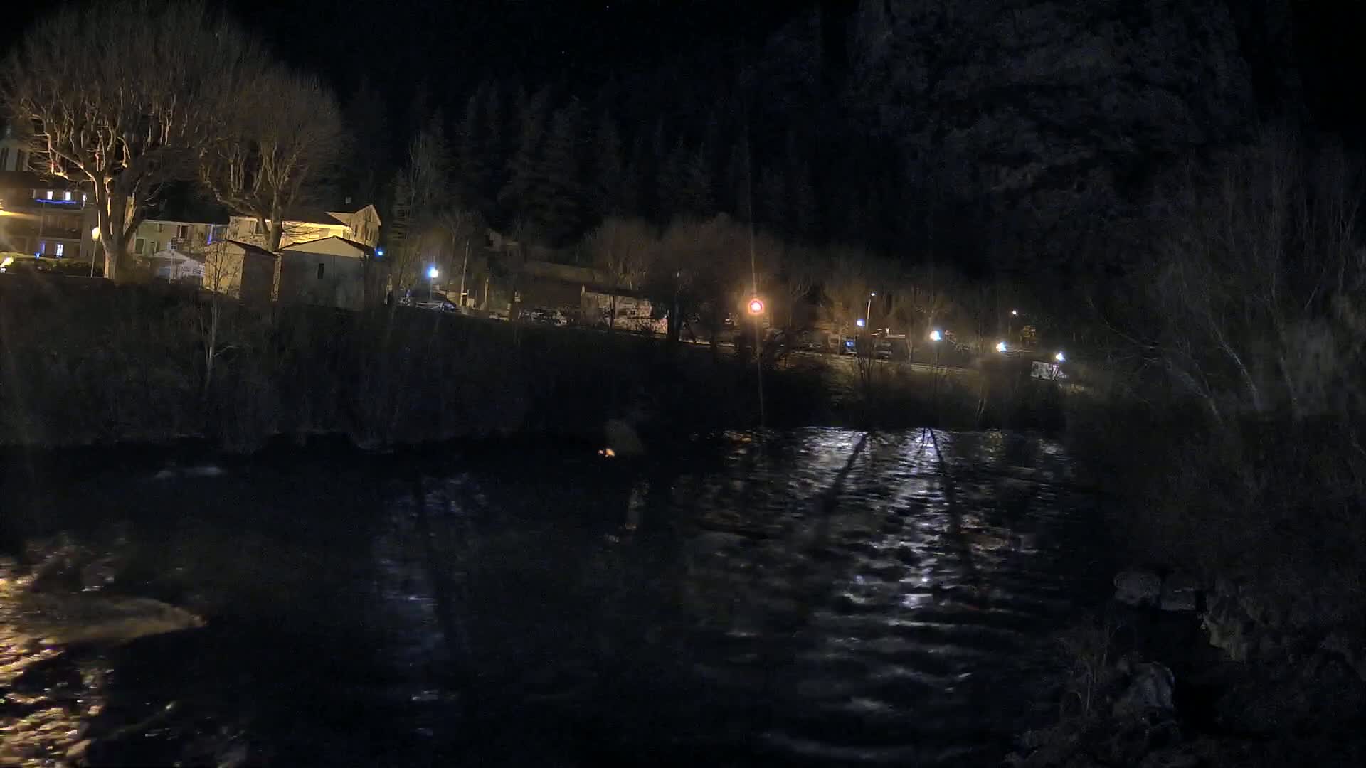 A dark, nighttime scene shows a river reflecting the lights of buildings and streetlights on the opposite bank, under a clear, starry sky.