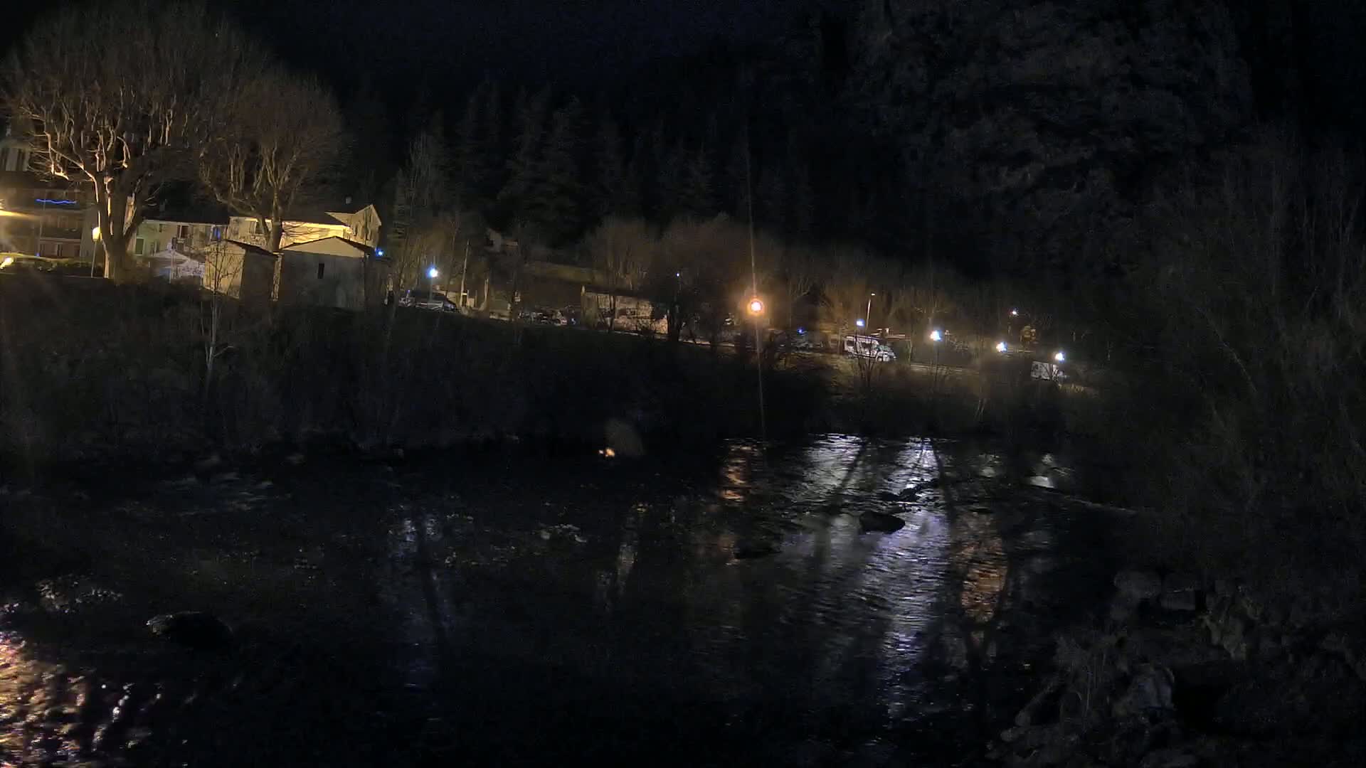 A dark river flows past a nighttime village, illuminated by streetlights and the faint glow of buildings.