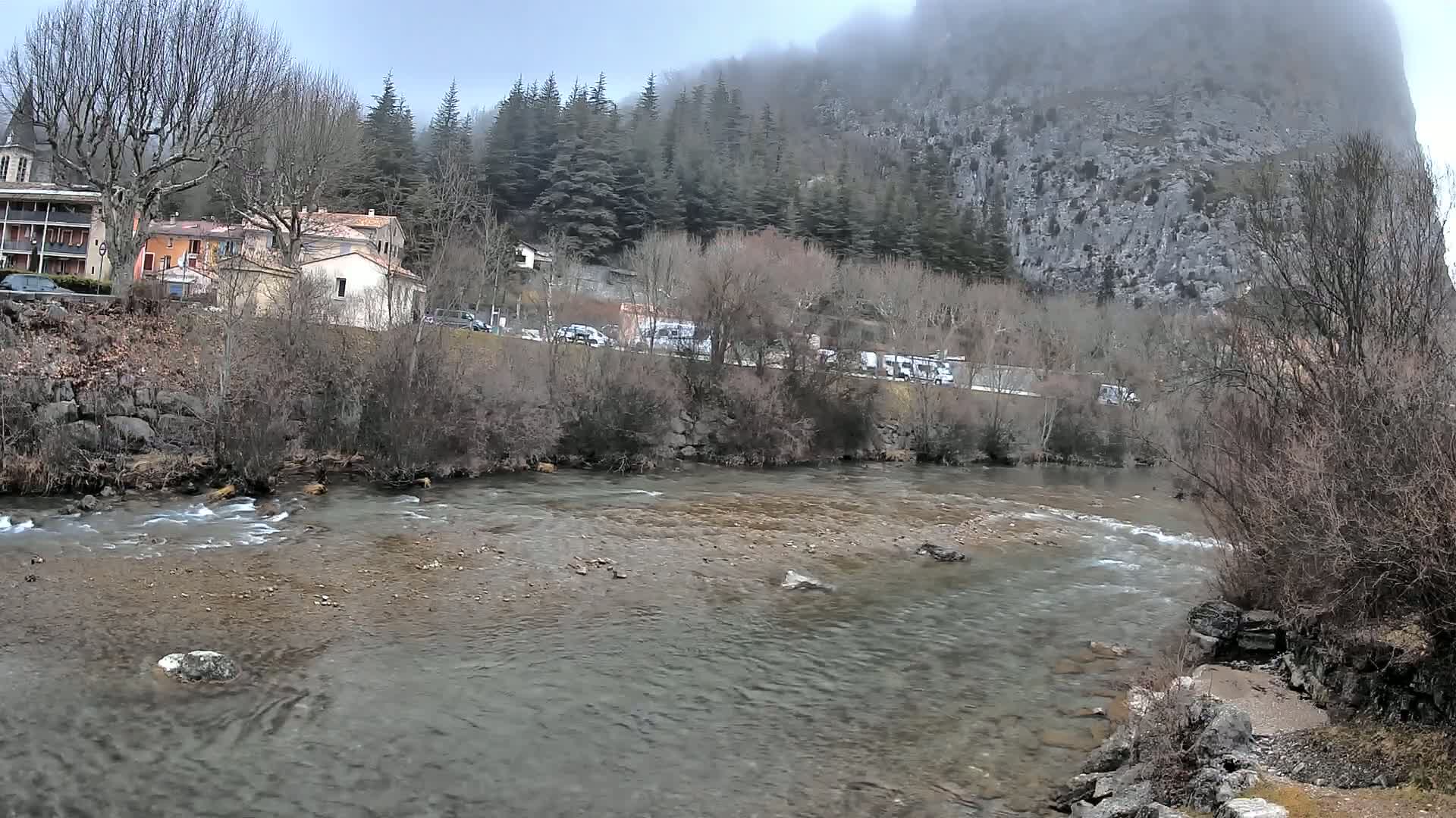 A slow-moving river flows past a village nestled at the base of a large, mist-covered mountain.
