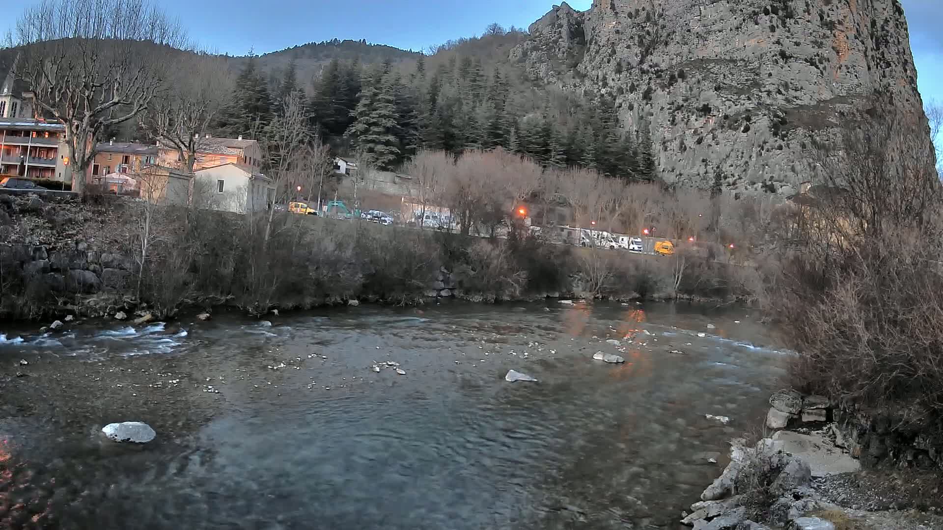 A river flows gently past buildings and a rocky cliff under a dusky sky.