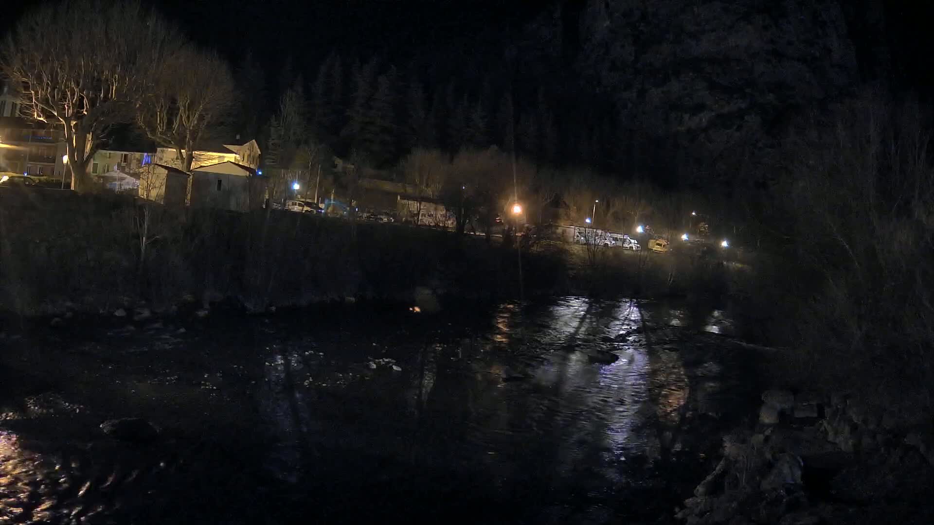A dark river flows past a small, nighttime village nestled among bare trees under a clear, black sky.