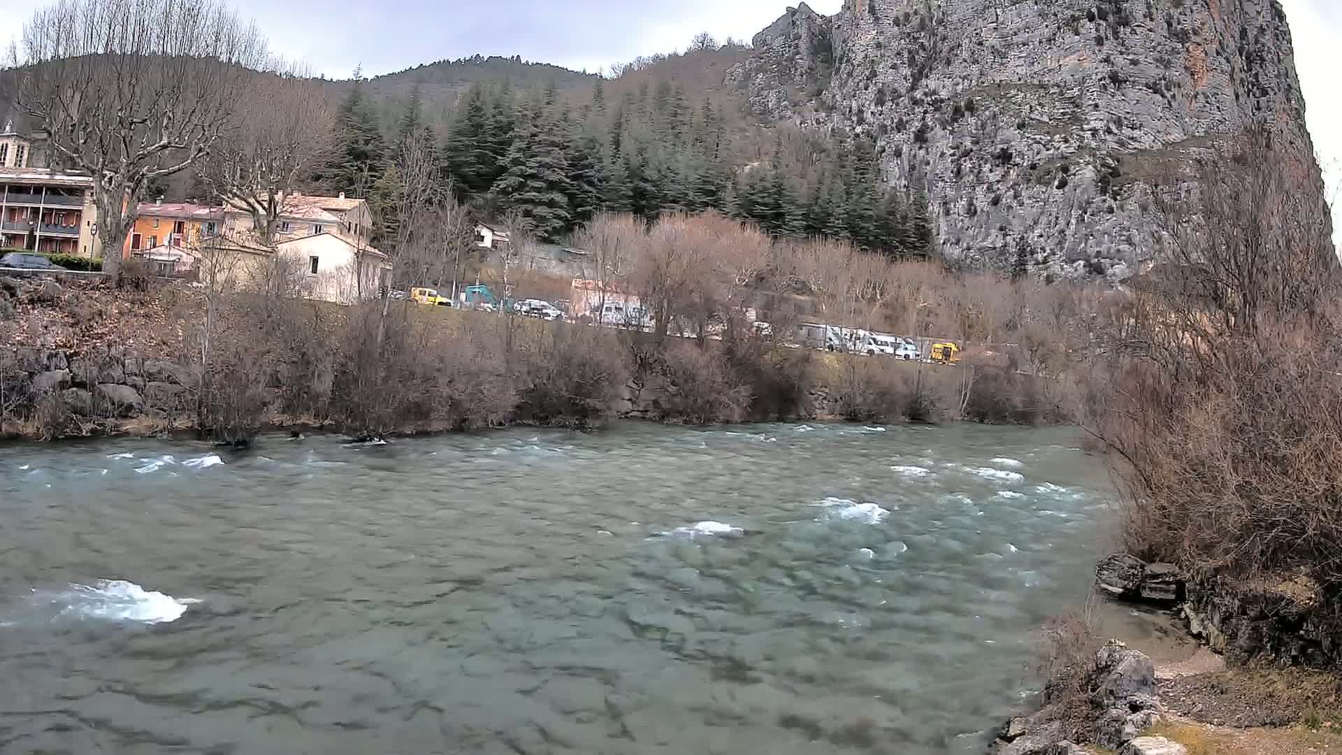 A swiftly flowing river runs in front of a village nestled beneath a large, grey cliff face on an overcast day.