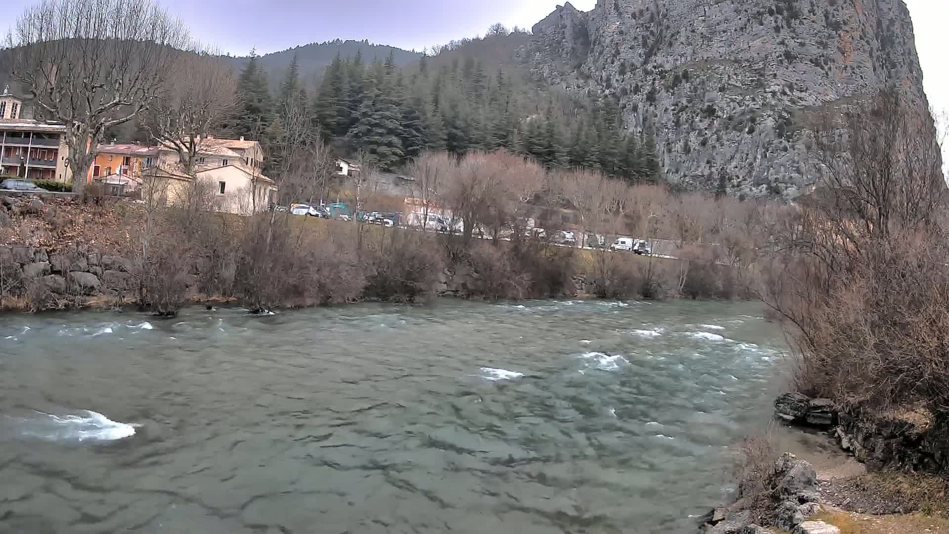 A swiftly flowing river runs past a village nestled beside a large, imposing rock face under an overcast sky.