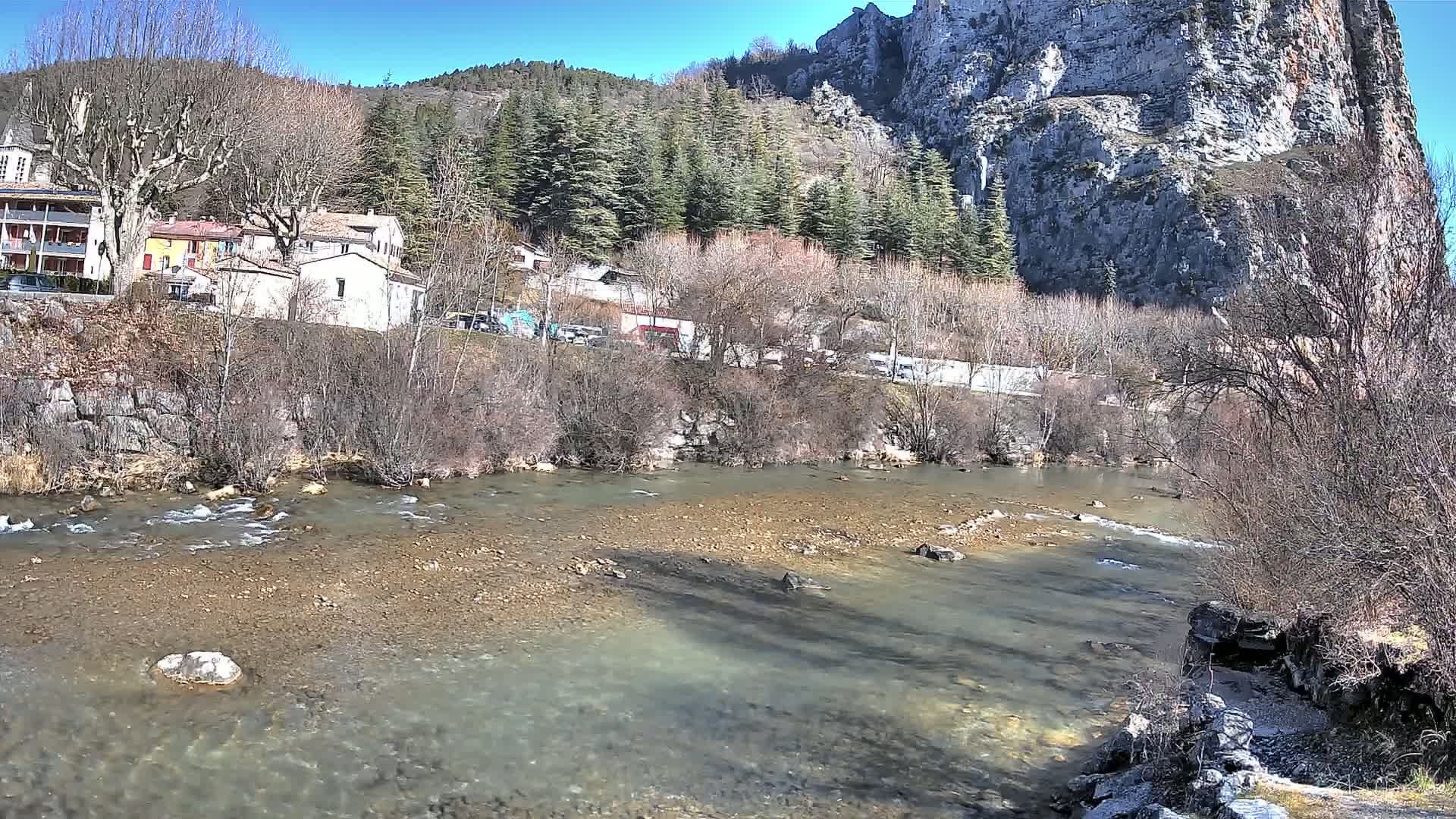A clear, shallow river flows past a village nestled beneath a large, rocky cliff face on a sunny day.