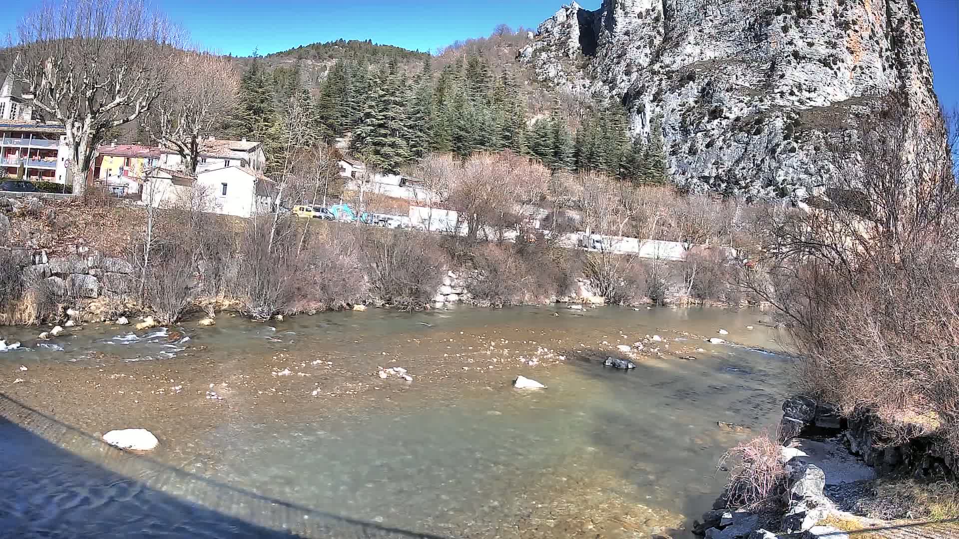 A clear, shallow river flows past a village nestled at the base of a large, rocky cliff under a sunny sky.