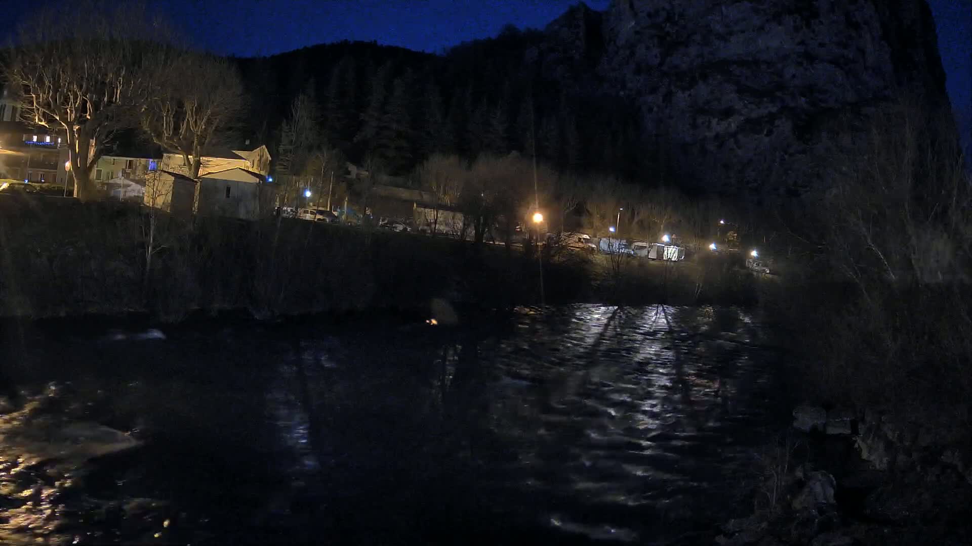 A dark, night scene shows a river flowing past a small village nestled at the base of a large, dark cliff under a clear, dark sky.