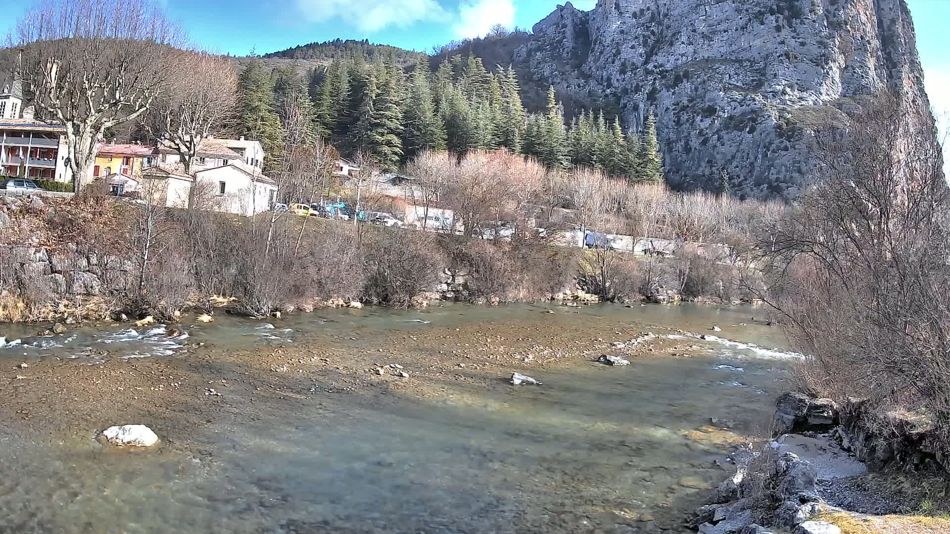A clear river flows past a village nestled at the base of a large, rocky cliff under a sunny sky.