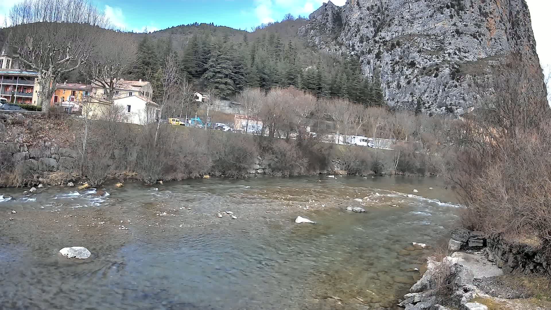 A clear river flows past a village nestled at the base of a rocky cliff under a partly cloudy sky.