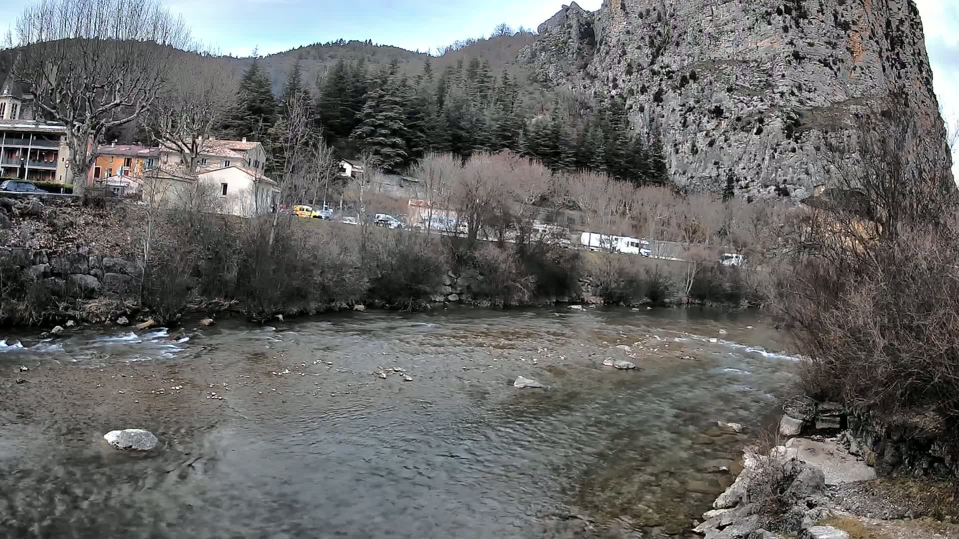 A river flows gently past a small village nestled at the base of a large, rocky cliff under a cloudy sky.