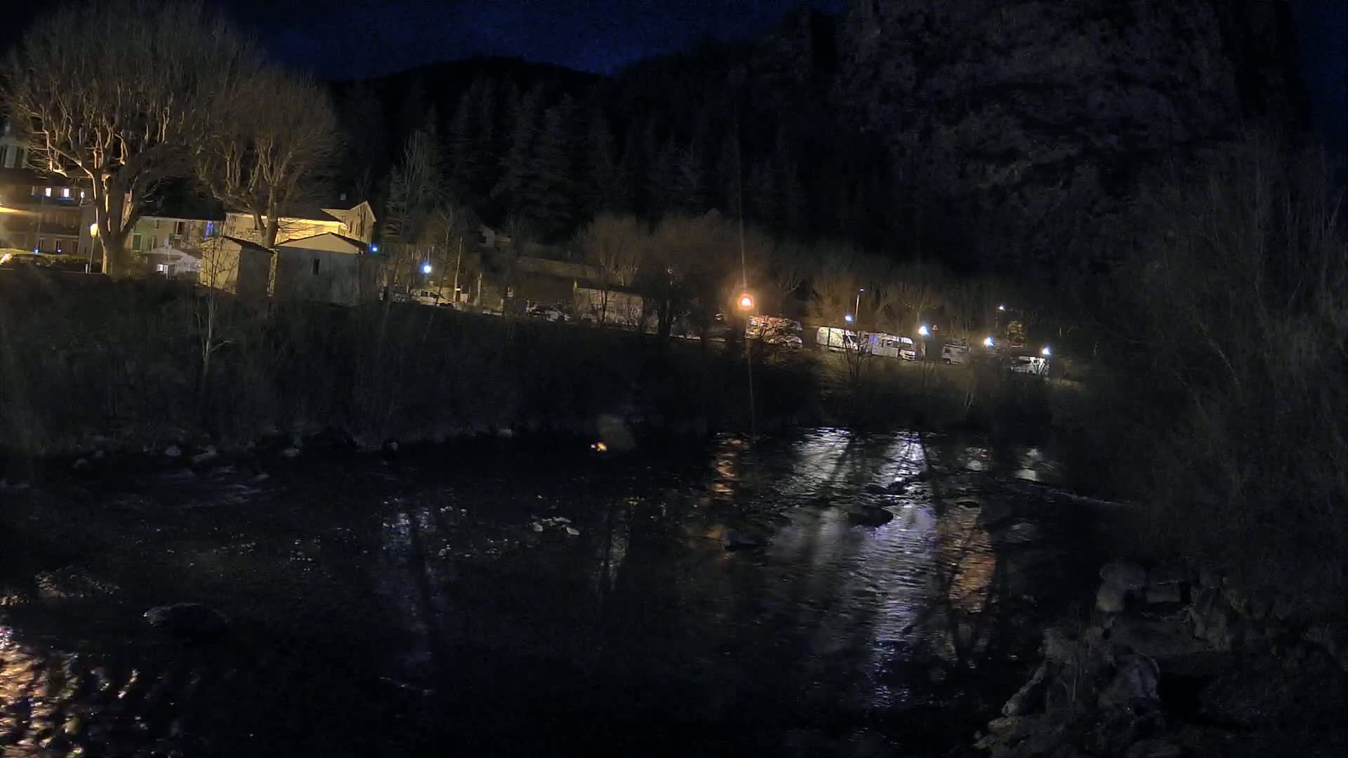 A dark, nighttime scene shows a river reflecting lights from buildings and vehicles along its bank, set against a backdrop of trees and hills.