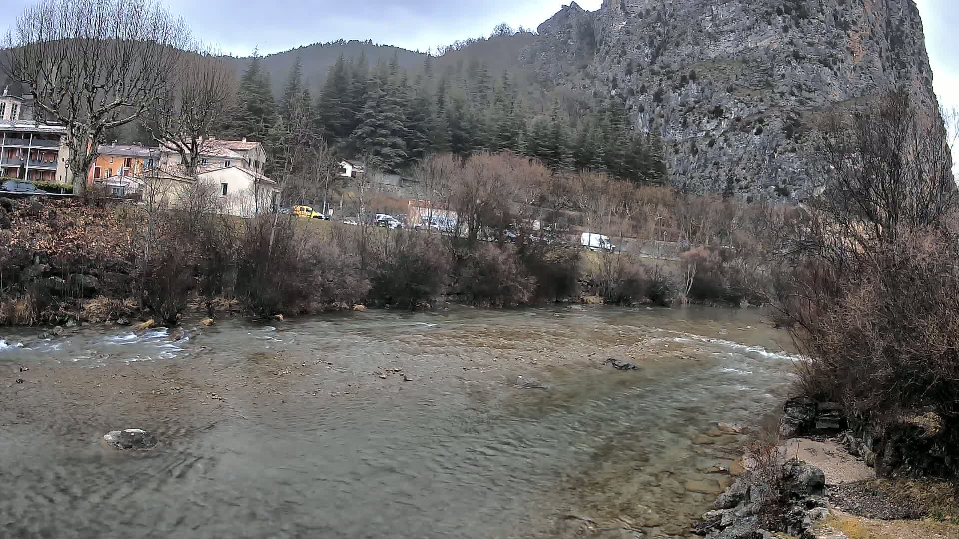 A slow-moving river flows past buildings and bare trees under an overcast sky, with a large rock face rising in the background.