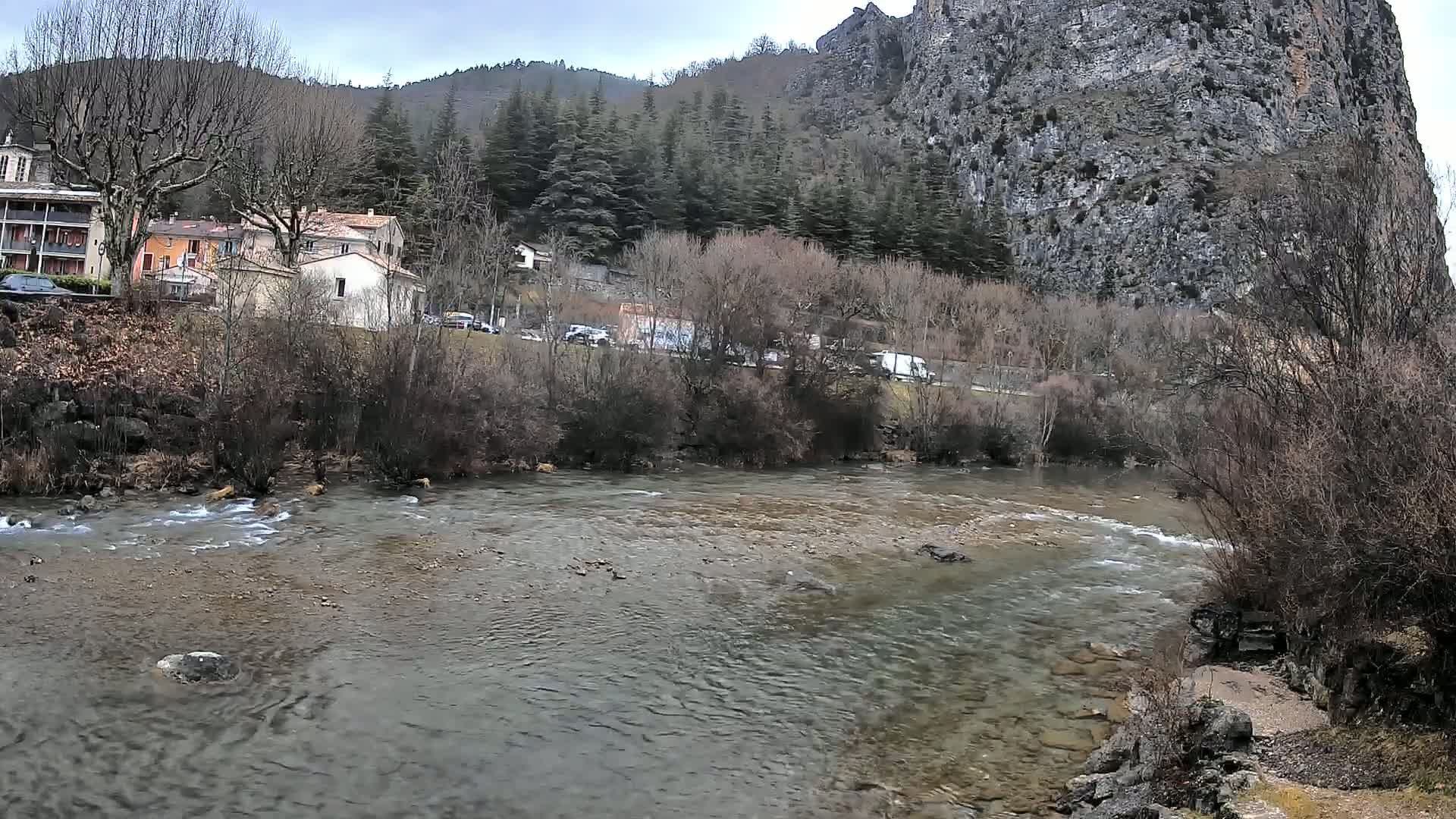 A river flows gently past a village nestled at the base of a large, grey cliff, under an overcast sky.