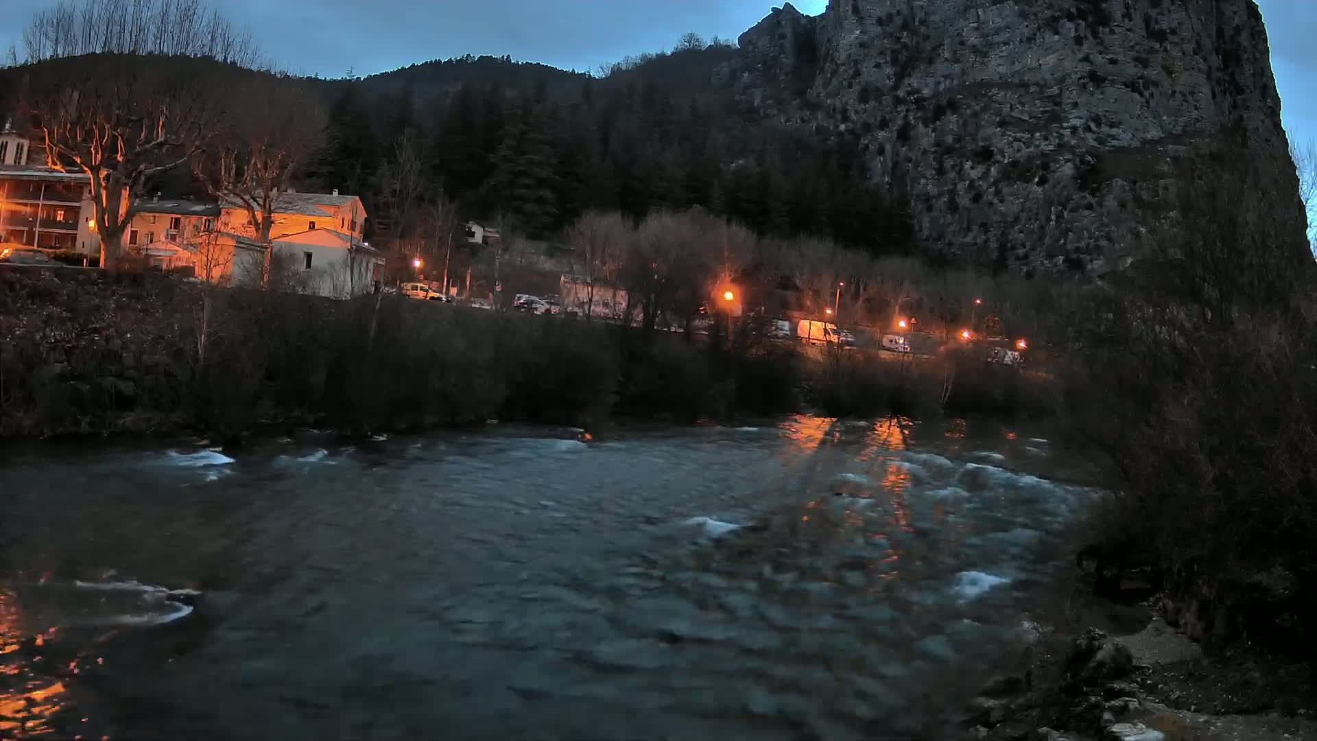 A river flows past a village nestled beside a large, dark cliff under a twilight sky.