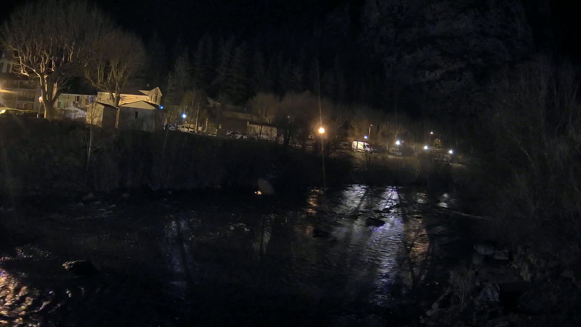 A dark river reflects the lights of a nighttime village nestled beside a dark forest.
