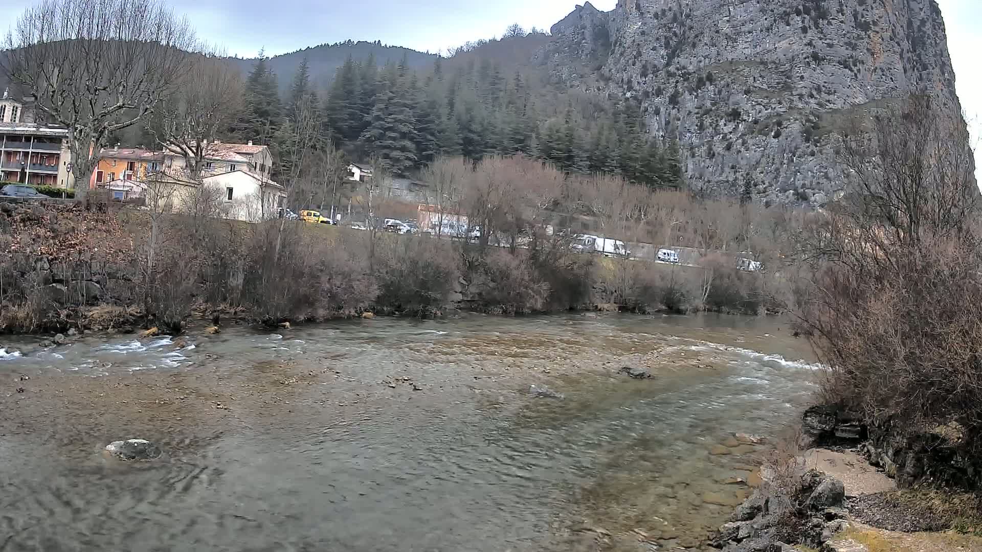 A river flows past a village nestled at the base of a large, rocky cliff, under an overcast sky.