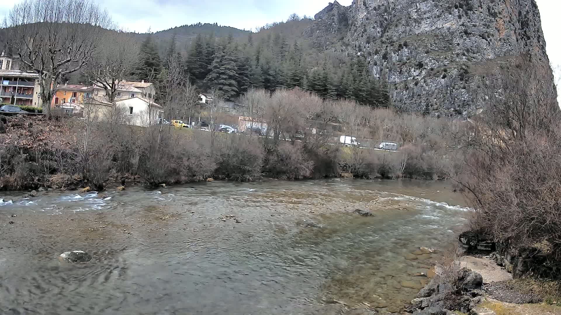 A rocky river flows past a small village nestled at the base of a steep, forested cliff on an overcast day.