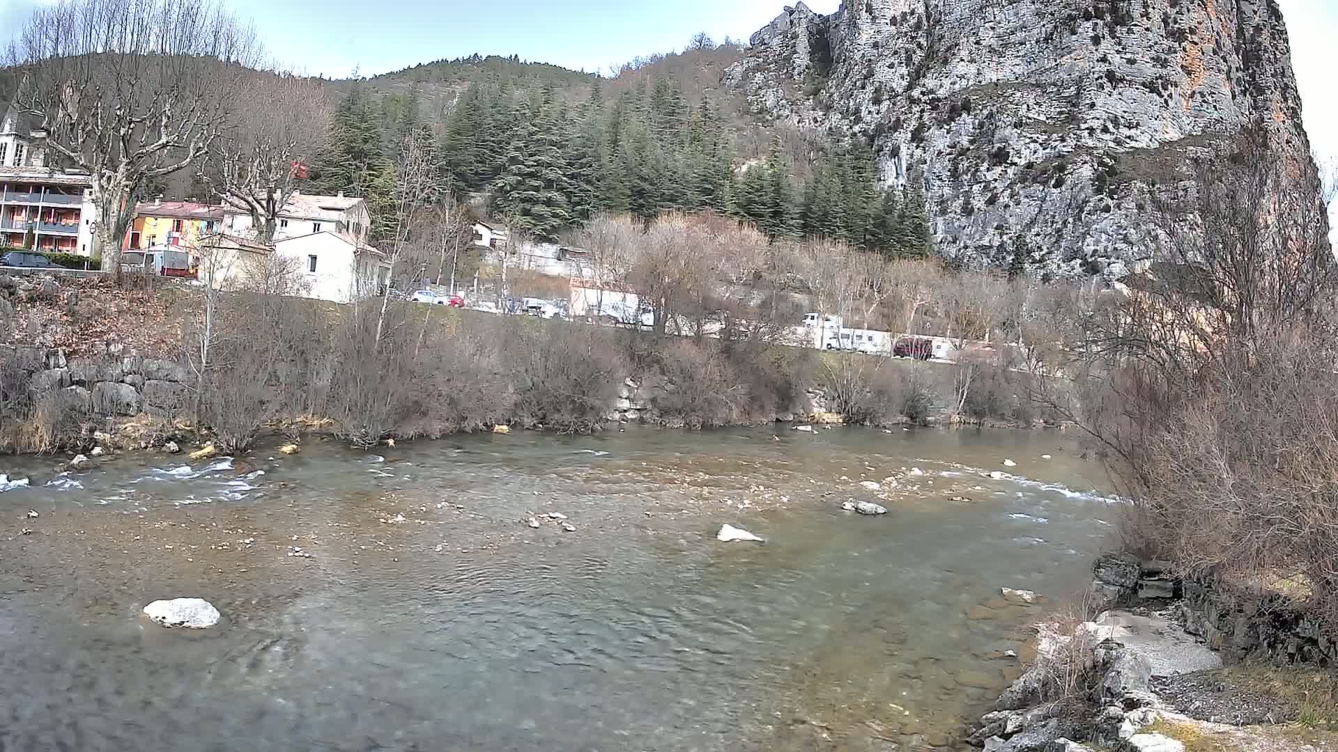 A clear river flows past a village nestled at the base of a large, rocky cliff under a partly cloudy sky.