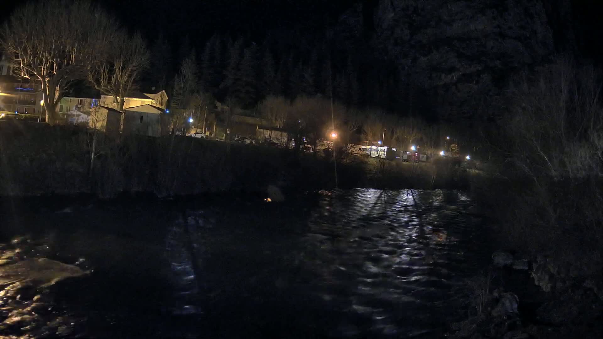 A dark, nighttime view of a flowing river beside a small town illuminated by scattered streetlights.