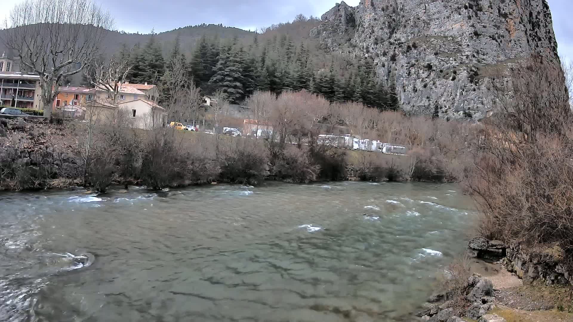 A river flows past a village nestled in a valley, under an overcast sky, with a large rock face rising behind the buildings and trees.