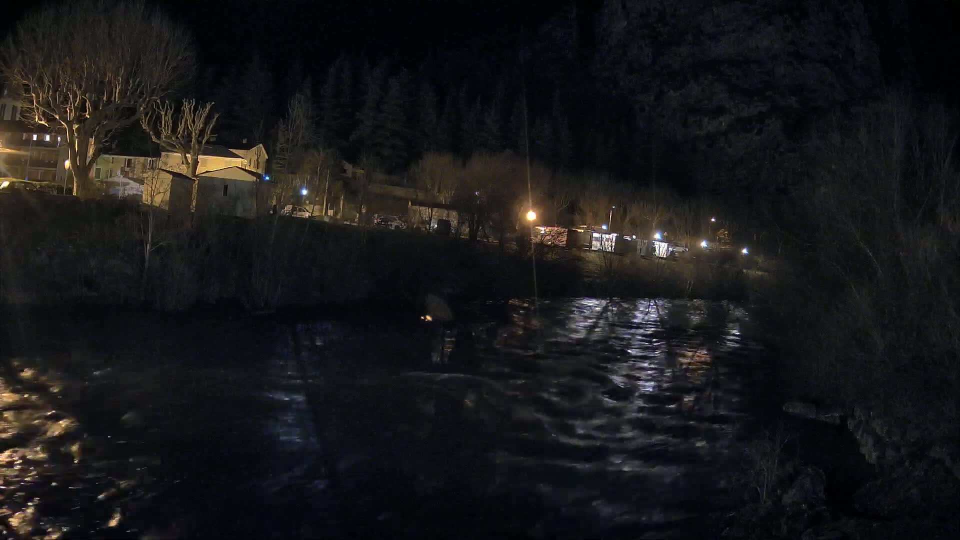 A dark, swiftly flowing river at night reflects the lights of a small village nestled against a hillside under a clear sky.