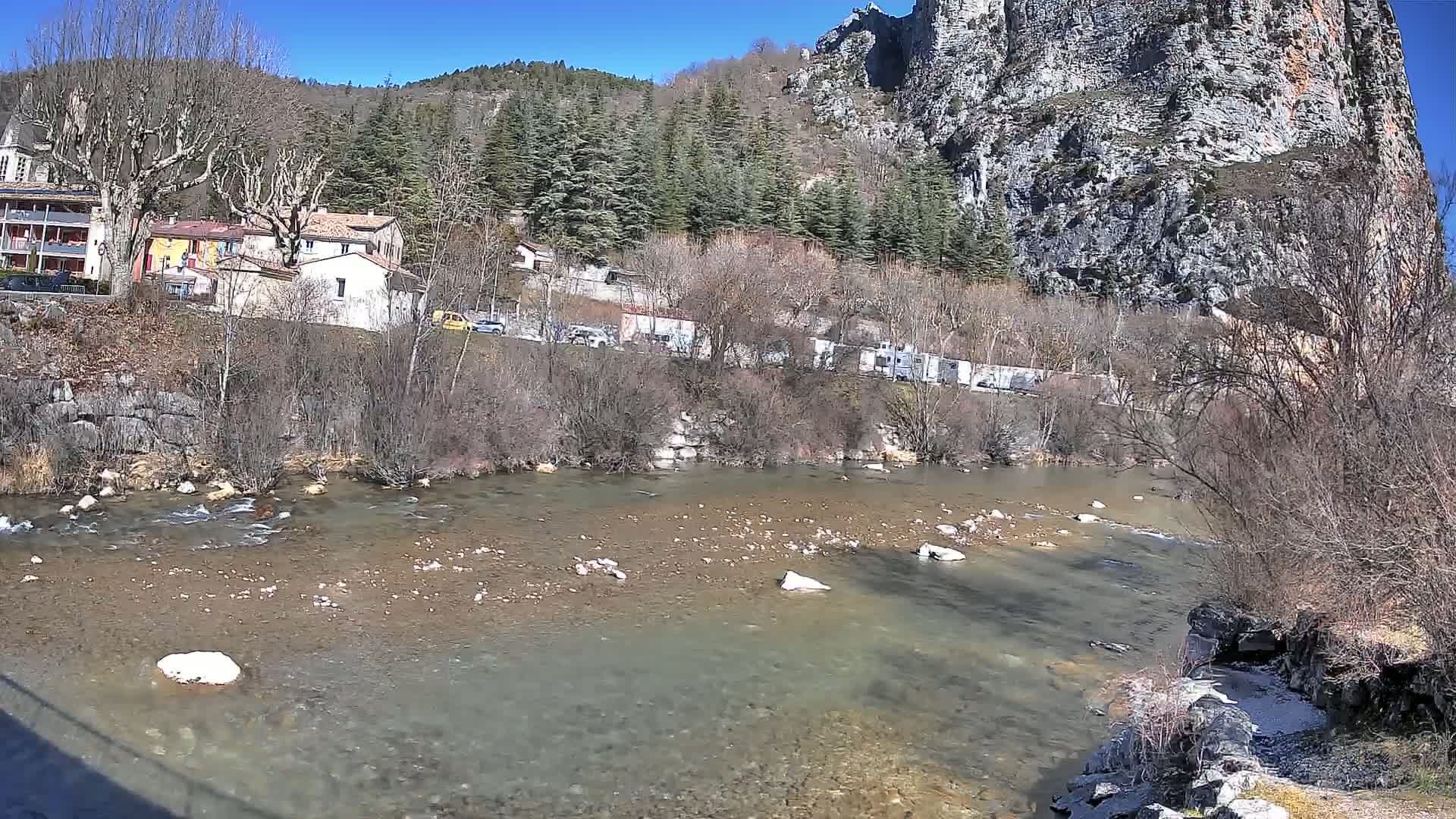 A clear, shallow river flows past a small village nestled at the base of a large, rocky cliff under a bright, sunny sky.