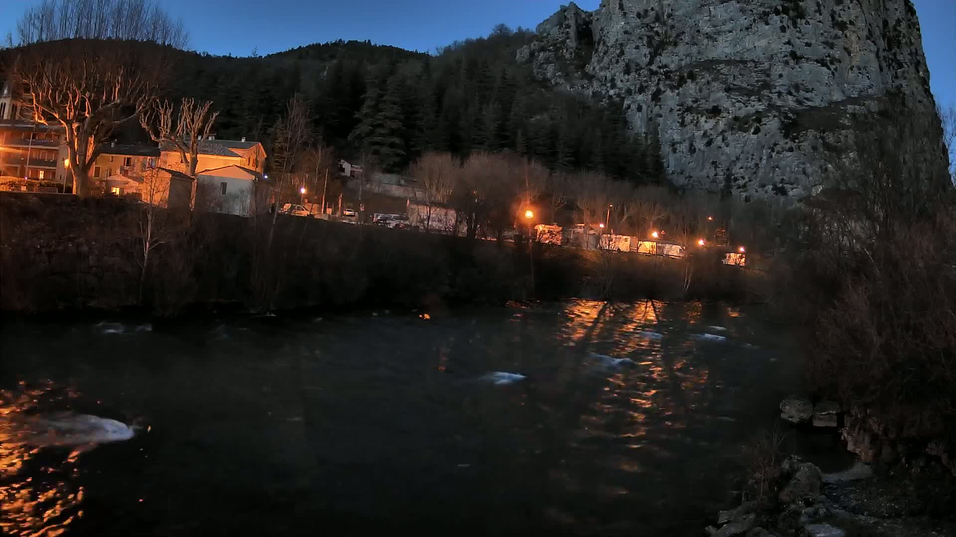 A river flows past a village nestled at the base of a large, dark cliff under a twilight sky.