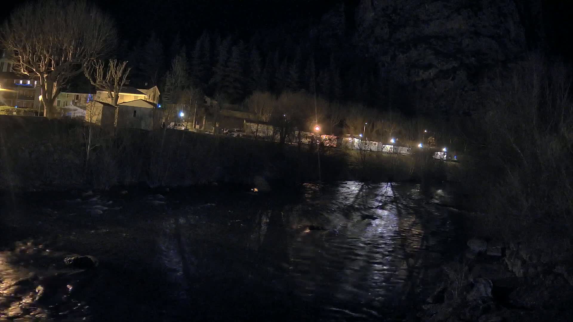 At night, a river flows past a dark, sparsely lit village nestled beside a wooded hillside.