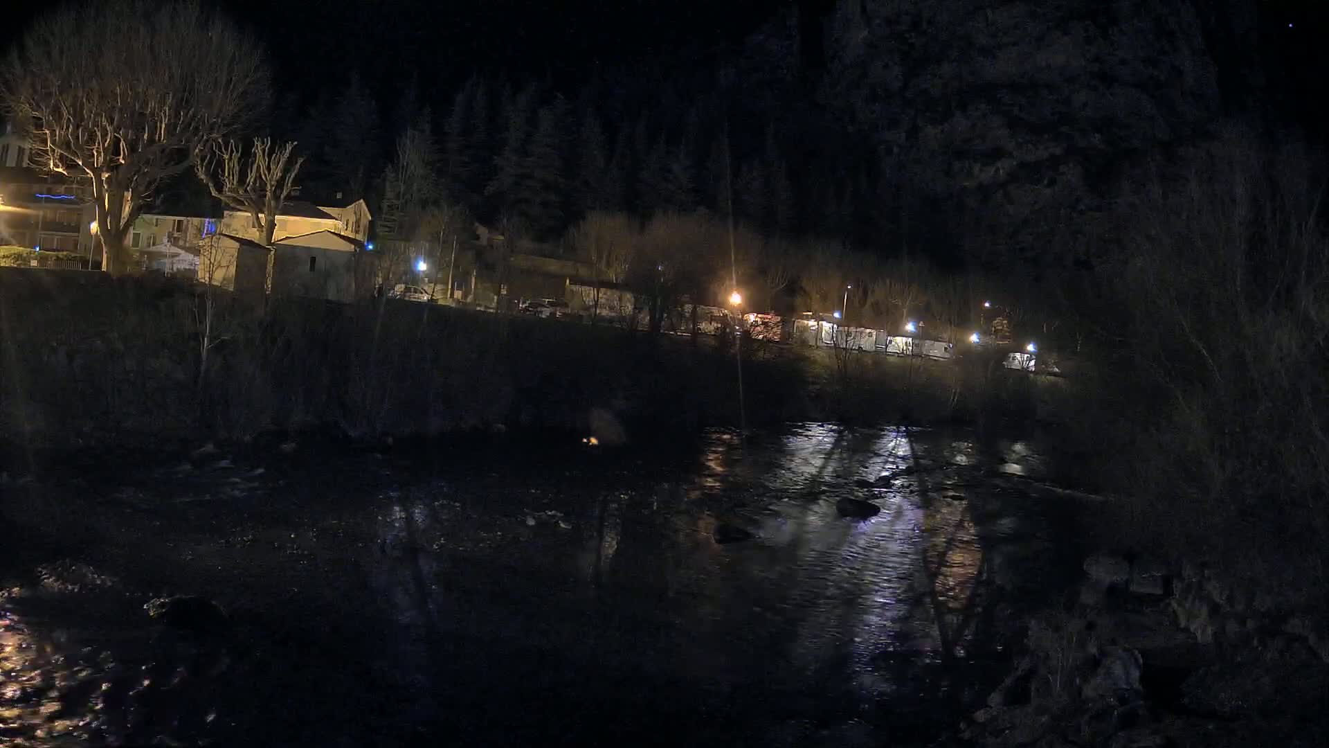 A dark, nighttime scene shows a river reflecting lights from buildings and vehicles on the opposite bank, under a clear sky.
