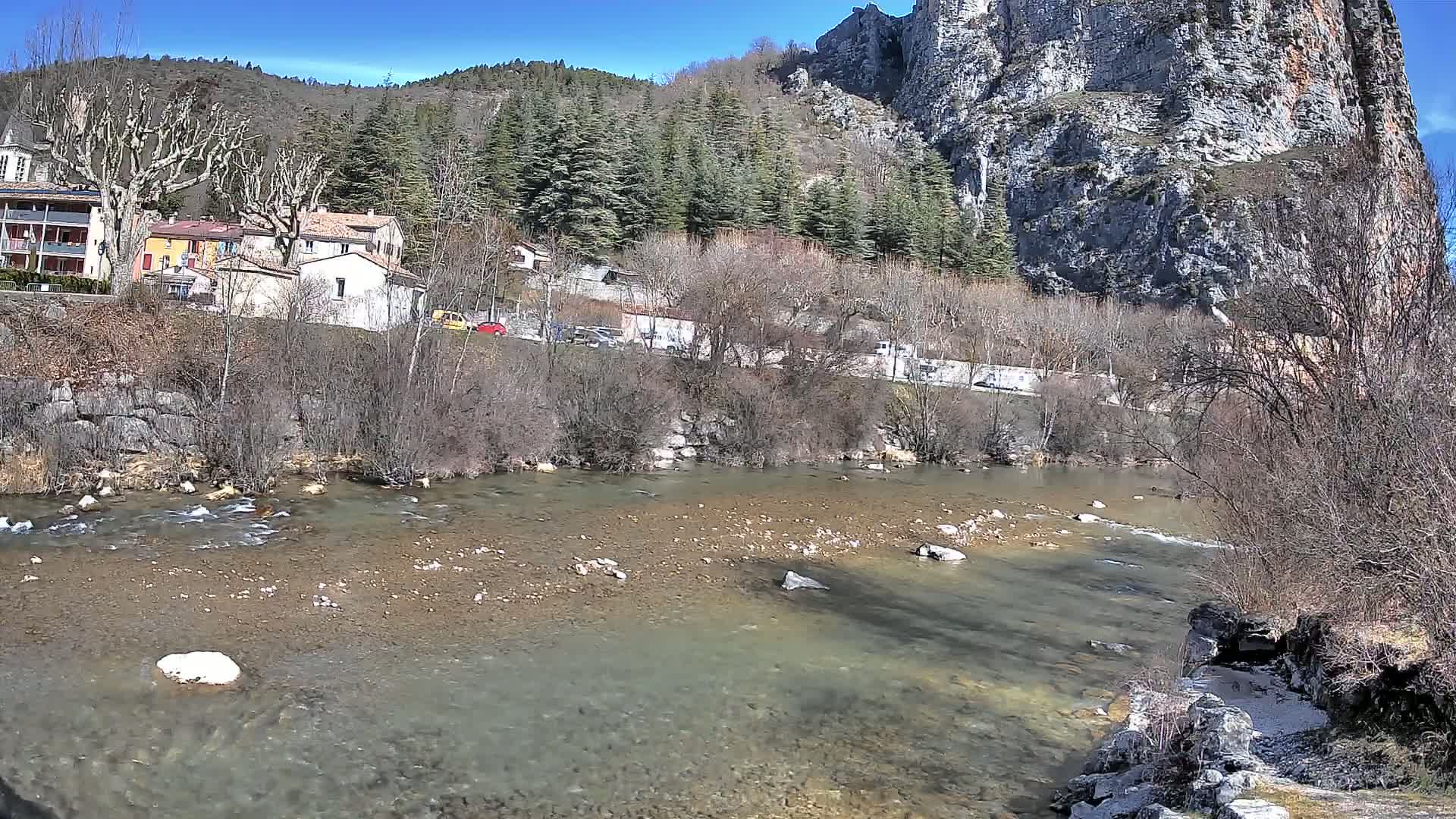 A shallow, clear river flows past a village nestled at the base of a large, rocky cliff under a sunny sky.